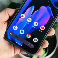 Dłoń trzyma smartfon z kolorową tapetą i ikonami aplikacji, w tym Sklep Play, Gmail, Zdjęcia i YouTube. Wygląda jak nowy android 15.