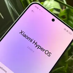 Ekran telefonu z napisem "Xiaomi HyperOS" w pastelowych barwach, otoczony zielenią roślin.