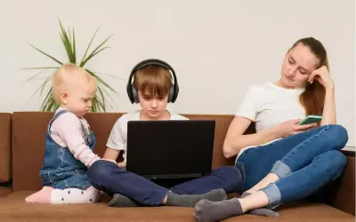 Dzieci na kanapie, chłopiec w słuchawkach gra na laptopie, maluch obok. Matka z telefonem. Kontrola rodzicielska w Polsce trwa do 18 lat.
