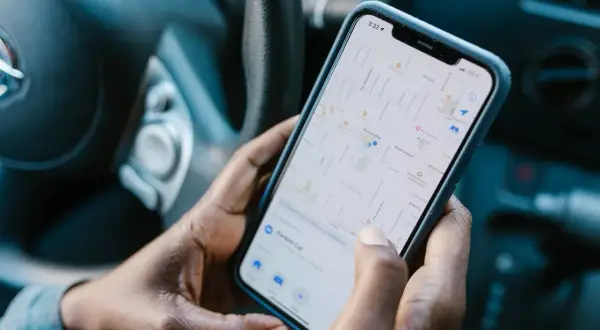 Dłonie trzymają telefon z mapą, pokazującą aplikację do udostępniania lokalizacji. Widać fragment kierownicy samochodu.