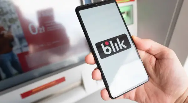 Dłoń trzyma smartfon z logo Blik. Obok bankomat, który ułatwia szybkie płatności. Kto wymyślił Blik? To polski system płatności.