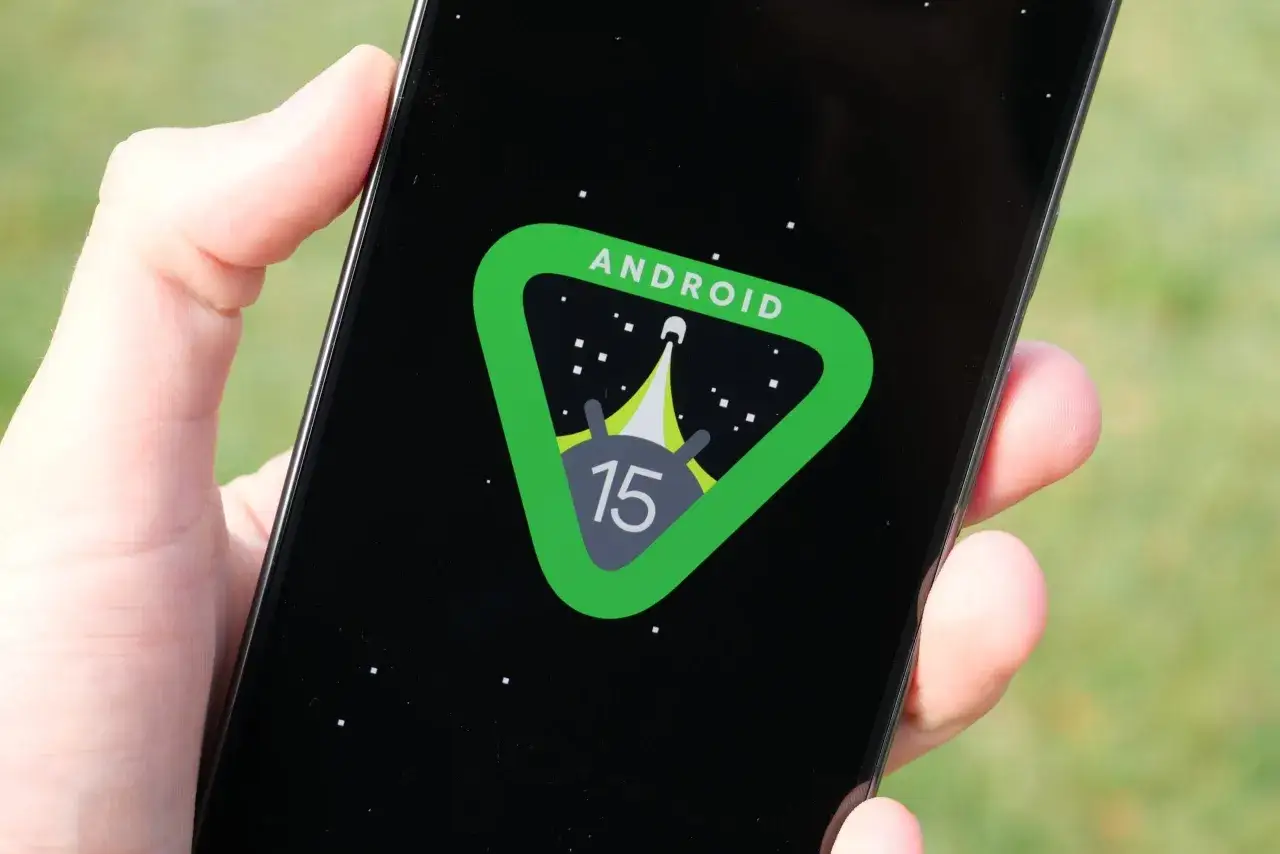Dłoń trzyma smartfon z logo Android 15 na czarnym tle, przypominającym kosmiczną podróż.