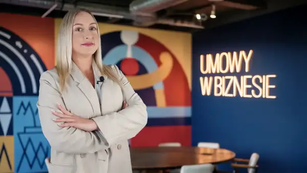 Kobieta w beżowej marynarce z mikrofonem, na tle neonu "UMOWY W BIZNESIE". Czy vider jest bezpieczny?