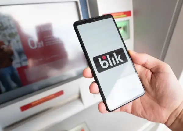 Dłoń trzyma smartfon z logo Blik. Obok bankomat, który ułatwia szybkie płatności. Kto wymyślił Blik? To polski system płatności.
